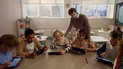 Six jeunes élèves munis de tablettes sont assis ensemble à une table de classe, et un enseignant regarde un exercice en cours par-dessus l'épaule d'un élève
