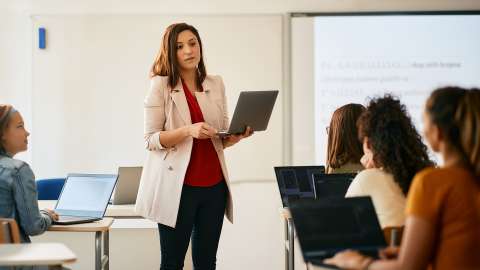 Enseignant tenant un PC portable ouvert devant une classe d'élèves, chacun ayant un PC portable ou un chromebook sur son bureau